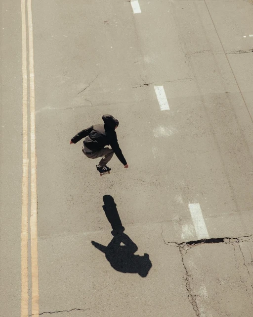 Young skateboarder in mid-air on an empty street, captured from an overhead angle, with the subject's shadow visible on the grey concrete.