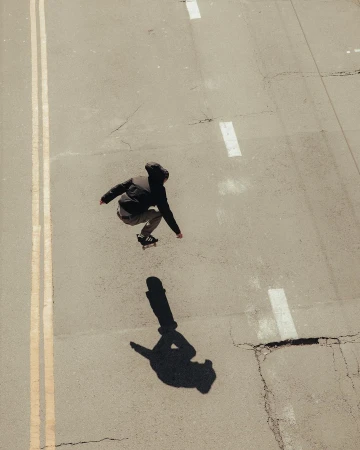 Young skateboarder in mid-air on an empty street, captured from an overhead angle, with the subject's shadow visible on the grey concrete.