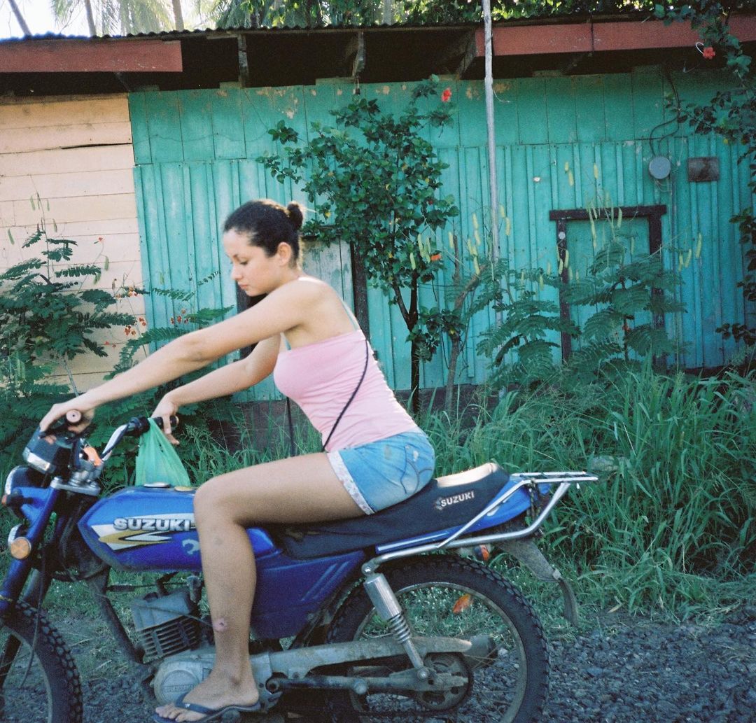 Young woman in pink top and blue denim shorts sits astride a blue Suzuki motorcycle on a rural road with a building in the background.