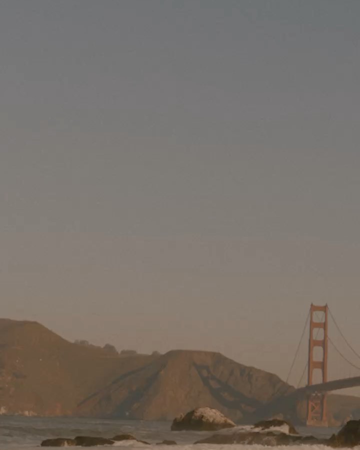 Golden Gate Bridge and surrounding hills during a hazy sunset with muted tones and a rocky shoreline in the foreground.