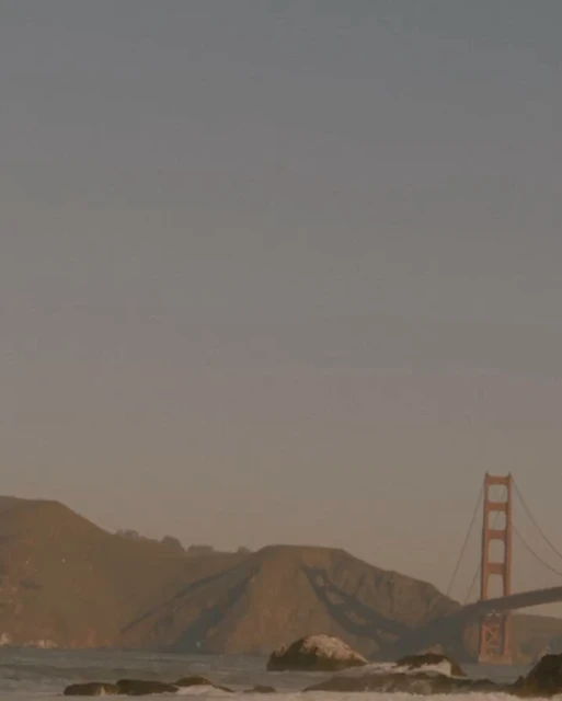 Golden Gate Bridge and surrounding hills during a hazy sunset with muted tones and a rocky shoreline in the foreground.