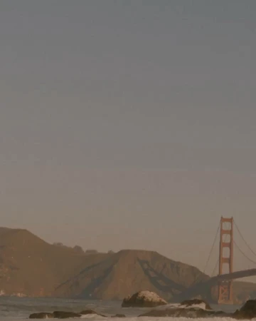 Golden Gate Bridge and surrounding hills during a hazy sunset with muted tones and a rocky shoreline in the foreground.