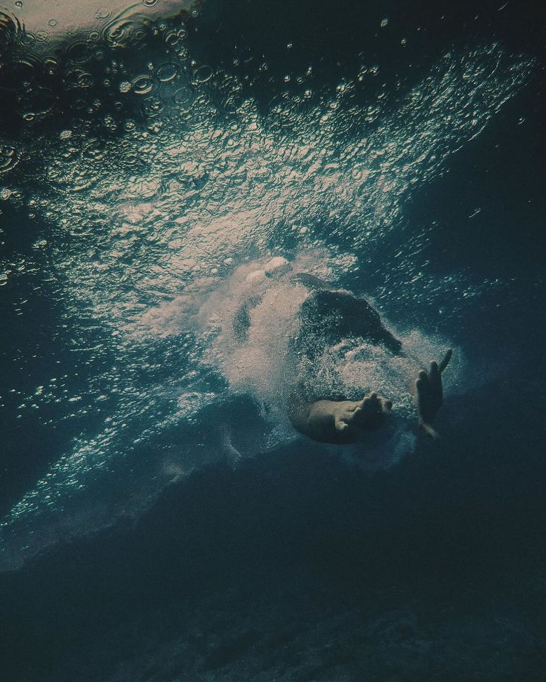 Underwater shot of a person swimming in deep, blue water with sunlight filtering through the surface, creating bubbles and a sense of depth.