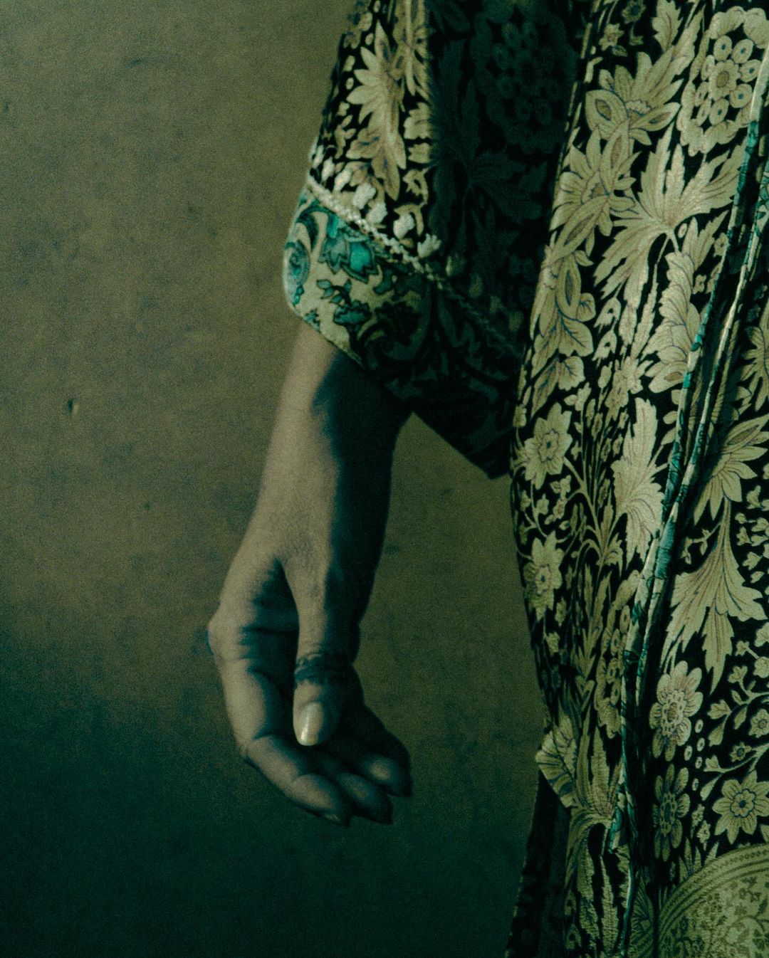 Close-up of an adult female's hand with a ring, contrasted against a green and black floral pattern robe and a neutral textured wall.