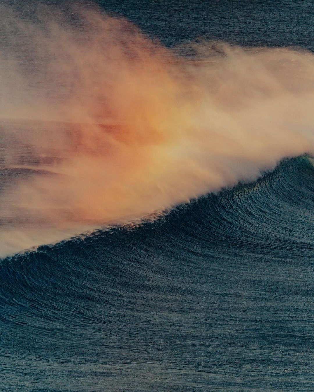 A powerful ocean wave curls toward the camera, its form blurred with motion, beneath a sky streaked with orange and purple hues.