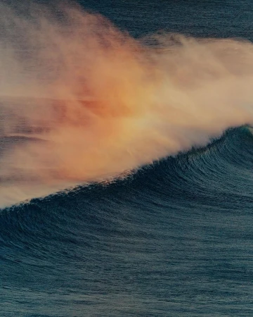 A powerful ocean wave curls toward the camera, its form blurred with motion, beneath a sky streaked with orange and purple hues.