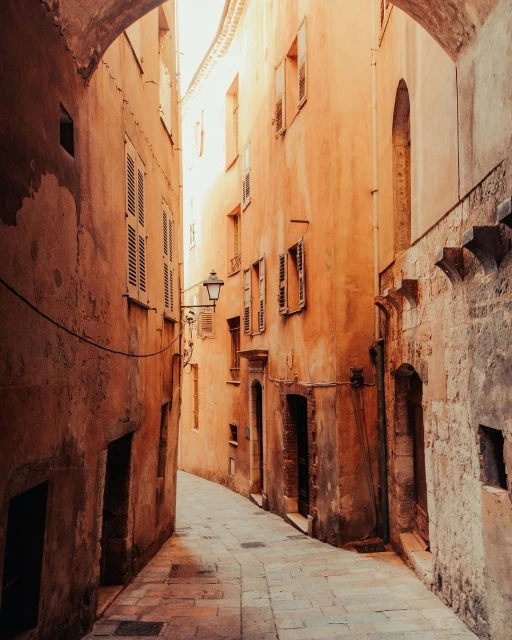 Narrow cobblestone street view between old orange buildings with weathered shutters, lit by sunlight and arching stone structures above.