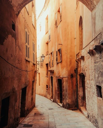 Narrow cobblestone street view between old orange buildings with weathered shutters, lit by sunlight and arching stone structures above.