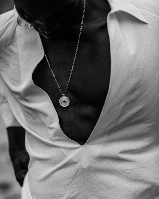 Black and white chest-level studio portrait of adult male in low-cut shirt with pendant necklace in a vertical composition.
