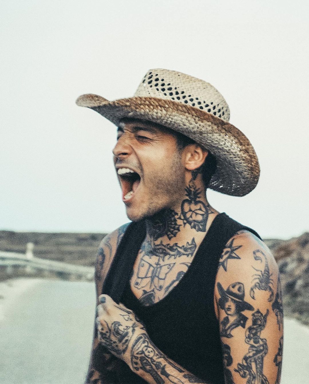 A man with tattoos and a cowboy hat yells with his mouth open on an open road with a white sky, wearing a black tank top.