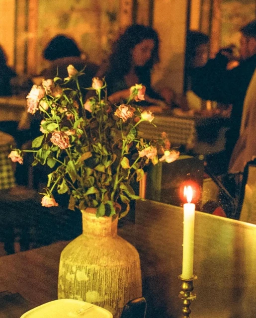 Still life of flowers in a vase and a candle in a restaurant, with blurred figures in the background dining at tables.