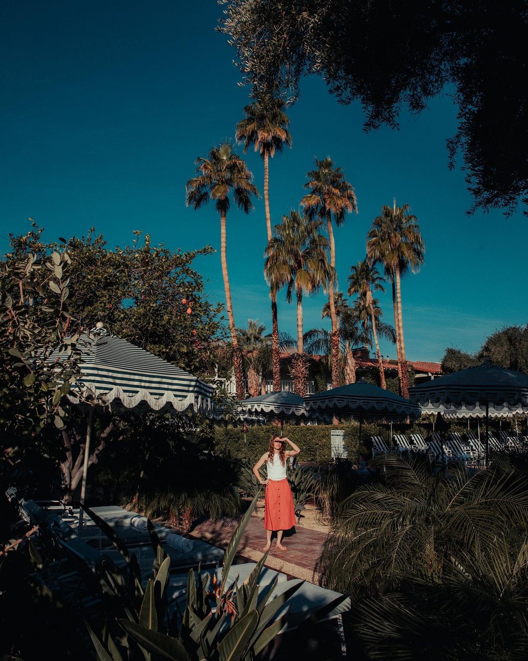 Adult female in red skirt stands on a stone pathway surrounded by tropical plants, palm trees and umbrellas on a sunny day.