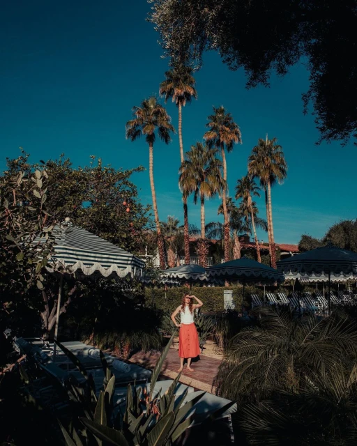 Adult female in red skirt stands on a stone pathway surrounded by tropical plants, palm trees and umbrellas on a sunny day.