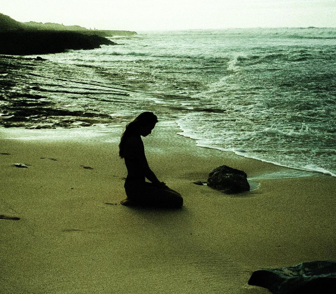 Silhouette of a woman kneeling on a sandy beach near ocean waves, with a rocky shoreline in the background, creating a serene coastal scene.