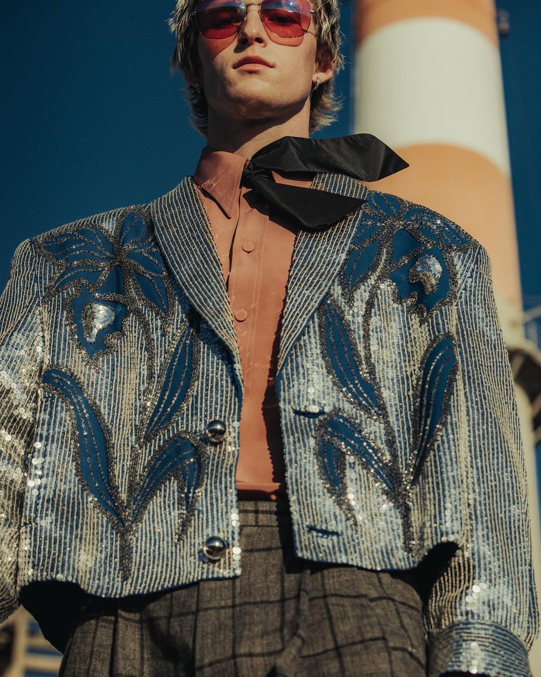 Young man in an embellished blue jacket, gray plaid pants, and pink sunglasses poses in front of a red and white industrial tower.