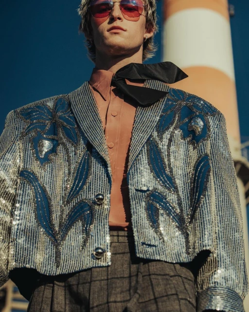 Young man in an embellished blue jacket, gray plaid pants, and pink sunglasses poses in front of a red and white industrial tower.