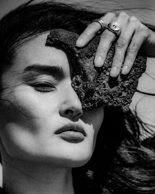Monochrome portrait of an adult female model with dark hair, light skin, and rings posing with a textured rock against her face.