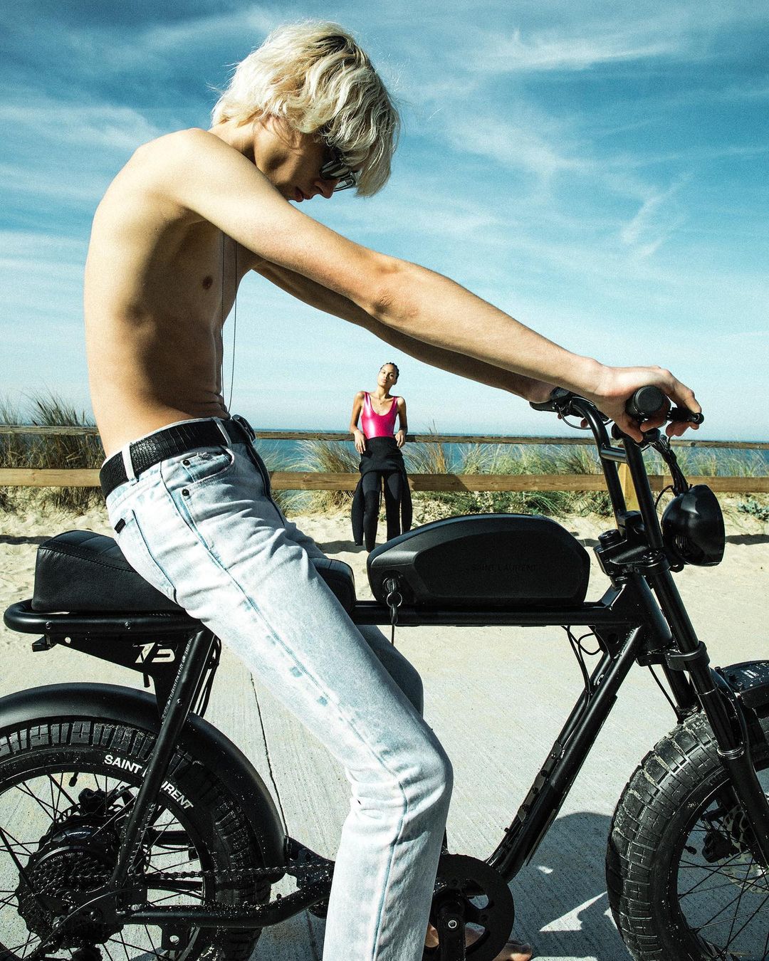 Young adult rides a black electric bike on a sandy beach under a bright sky, with another person standing near the ocean.