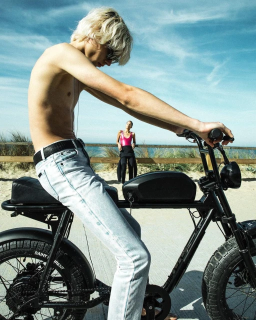 Young adult rides a black electric bike on a sandy beach under a bright sky, with another person standing near the ocean.