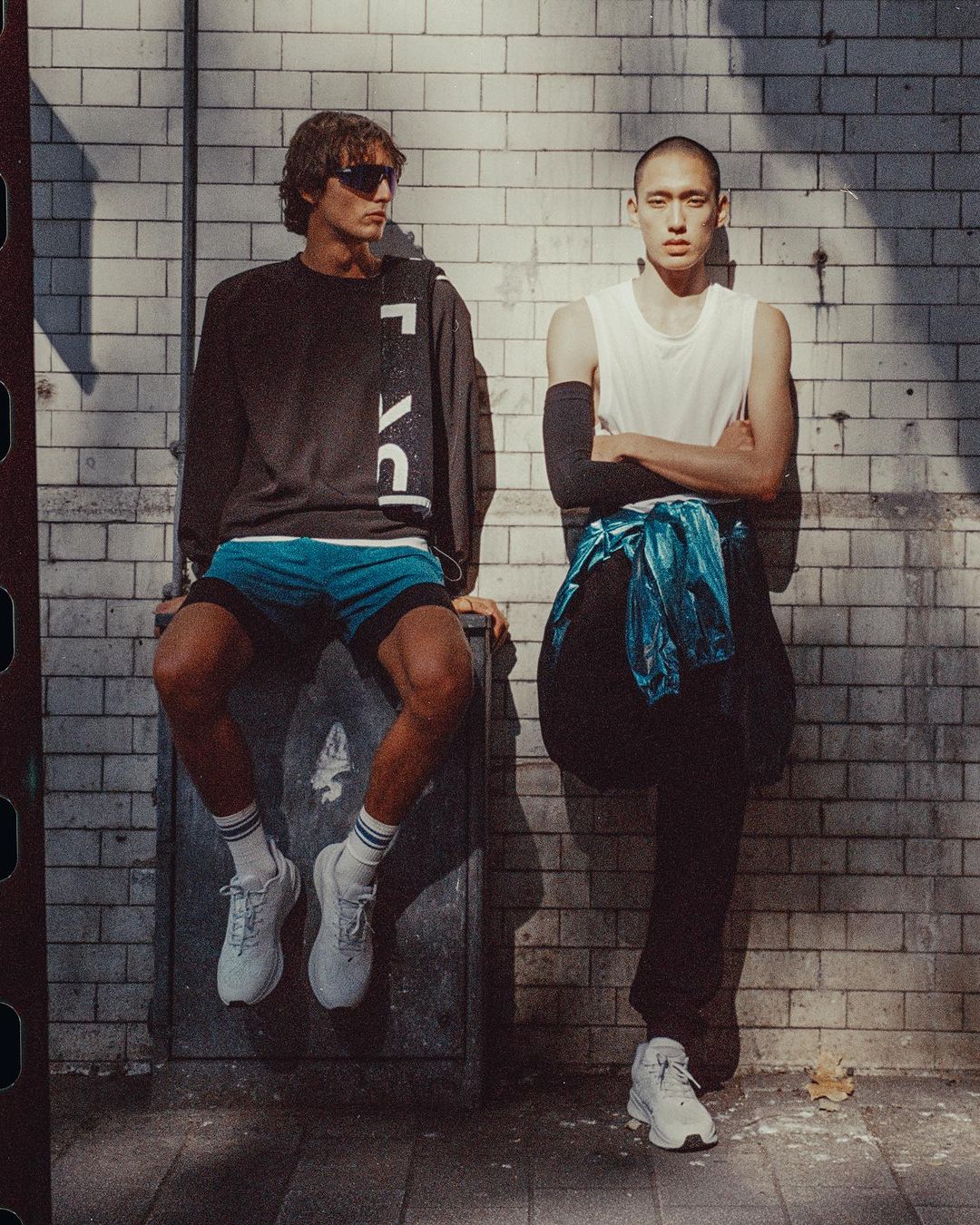 Two young men in athletic wear pose outdoors against a brick wall; one sits on a metal box, both wearing athletic shoes. Shadowy, bright lighting.