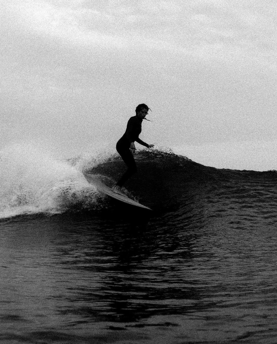 Black and white shot of a female surfer in a wetsuit riding a wave on a surfboard, with water splashing and a cloudy sky overhead.