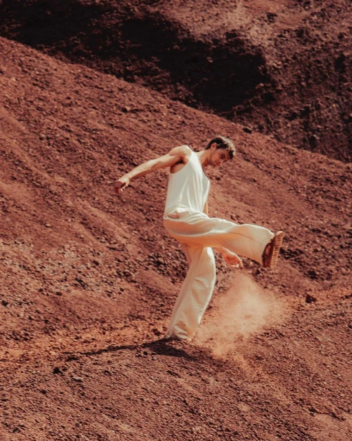 Adult male in a white tank top and pants kicks up red dirt on a hillside in an outdoor setting, facing towards the right.