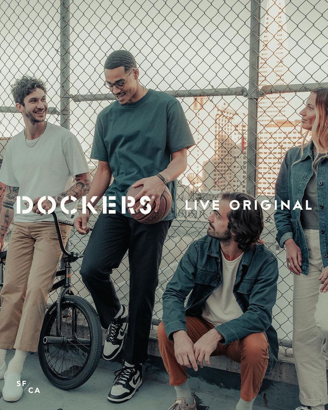 Five young adults gather outdoors in casual clothing, including t-shirts, jeans, and sneakers. One man holds a basketball near a chainlink fence.