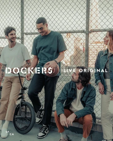 Five young adults gather outdoors in casual clothing, including t-shirts, jeans, and sneakers. One man holds a basketball near a chainlink fence.