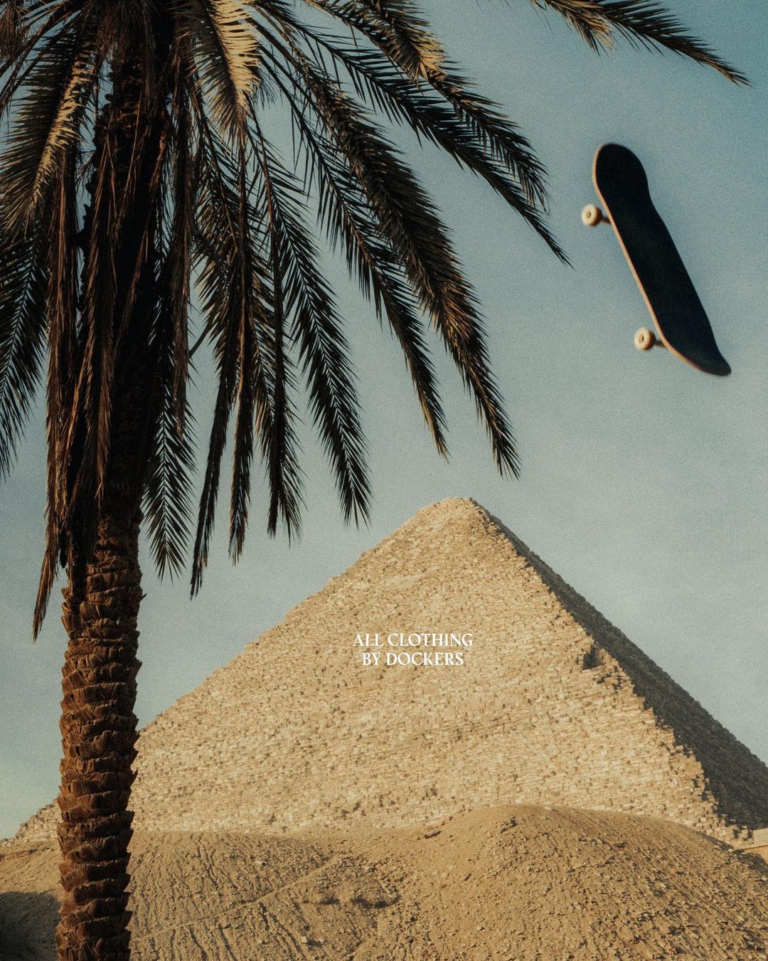 Low-angle shot of skateboard in mid-air near a palm tree and pyramid under a blue sky, showcasing urban and travel themes.