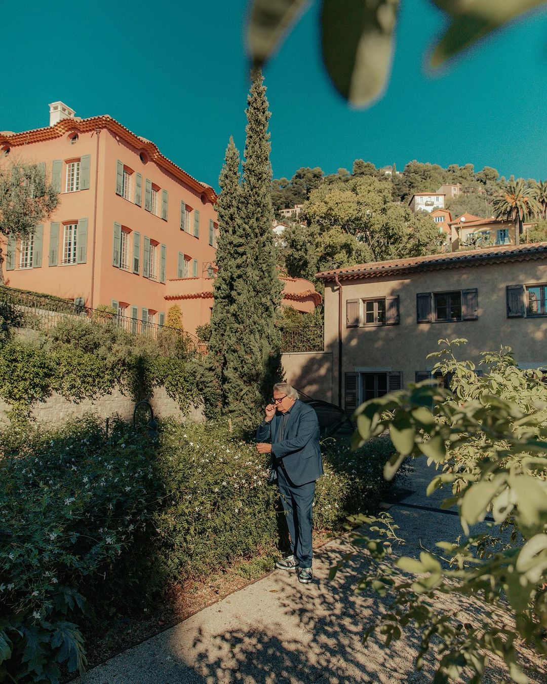 Adult male in a blue suit stands on a pathway in front of a villa with white flowers and green foliage in a rural outdoor setting.