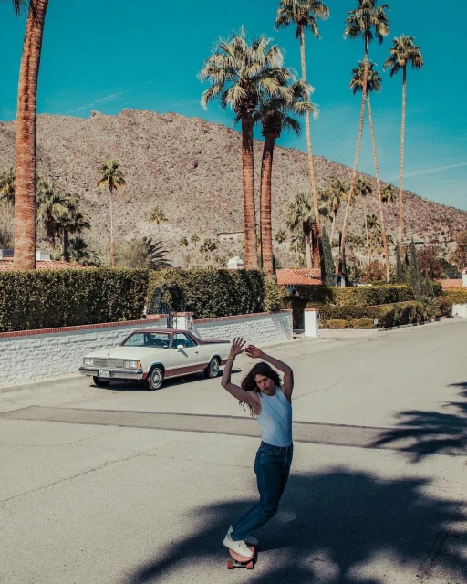 Adult female skateboarding on an asphalt road near palm trees and a vintage car under a clear blue sky with a mountain backdrop.