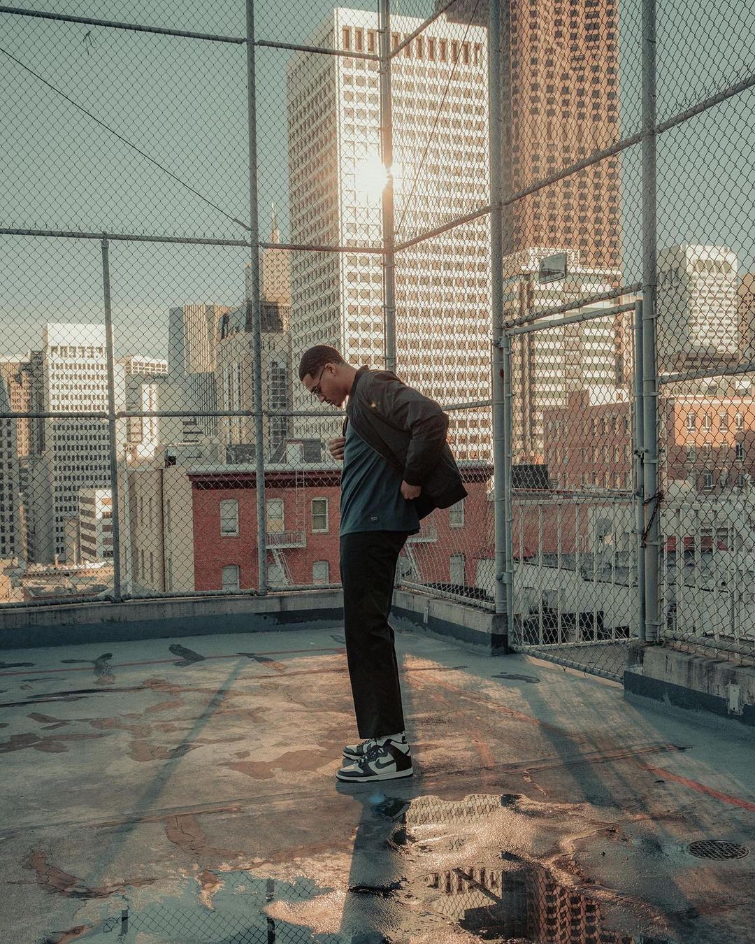 Man standing on a concrete rooftop behind a chain-link fence against a backdrop of city skyscrapers in sunlight.