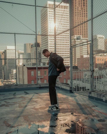 Man standing on a concrete rooftop behind a chain-link fence against a backdrop of city skyscrapers in sunlight.