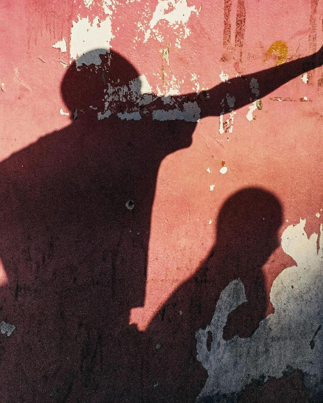Silhouette shadows of two people against a textured pink wall with weathered patches and peeled paint, creating an artistic contrast.