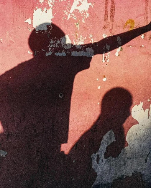 Silhouette shadows of two people against a textured pink wall with weathered patches and peeled paint, creating an artistic contrast.