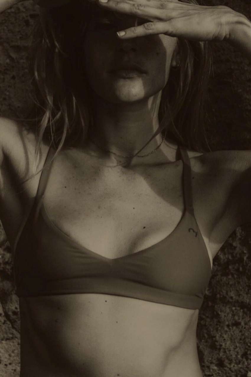 Woman in swimwear poses against a rock backdrop, hands shielding eyes. Shot in natural light with a warm, sepia-toned color palette.