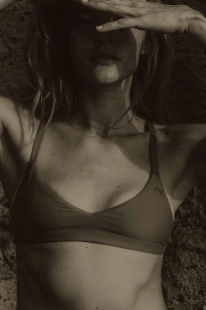 Woman in swimwear poses against a rock backdrop, hands shielding eyes. Shot in natural light with a warm, sepia-toned color palette.