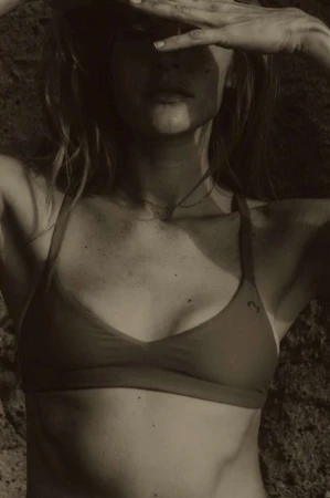 Woman in swimwear poses against a rock backdrop, hands shielding eyes. Shot in natural light with a warm, sepia-toned color palette.
