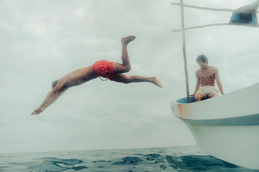 A man dives headfirst into the ocean from a small boat, with another man seated inside on an overcast day at sea.