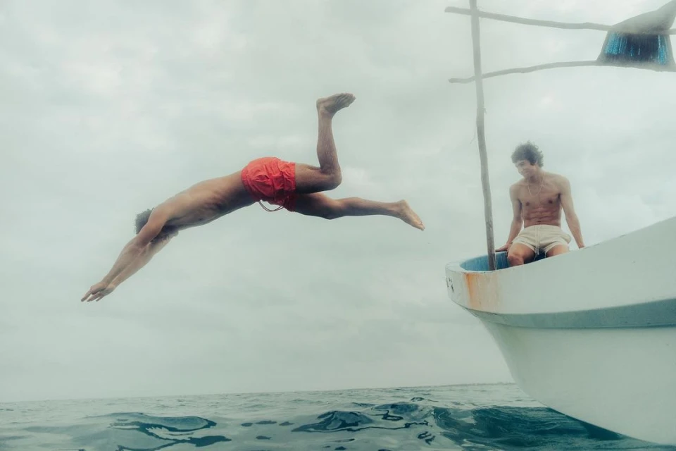 A man dives headfirst into the ocean from a small boat, with another man seated inside on an overcast day at sea.
