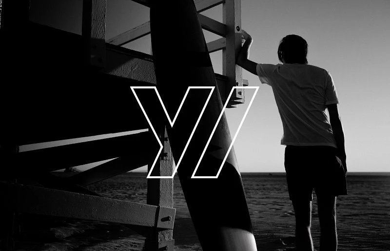 Monochrome shot of a person standing near a lifeguard tower on a beach under bright sunlight, with a logo overlay.