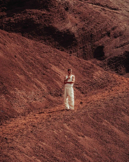Man in white outfit poses on a reddish-brown gravel hillside, looking to the left in a natural outdoor environment.