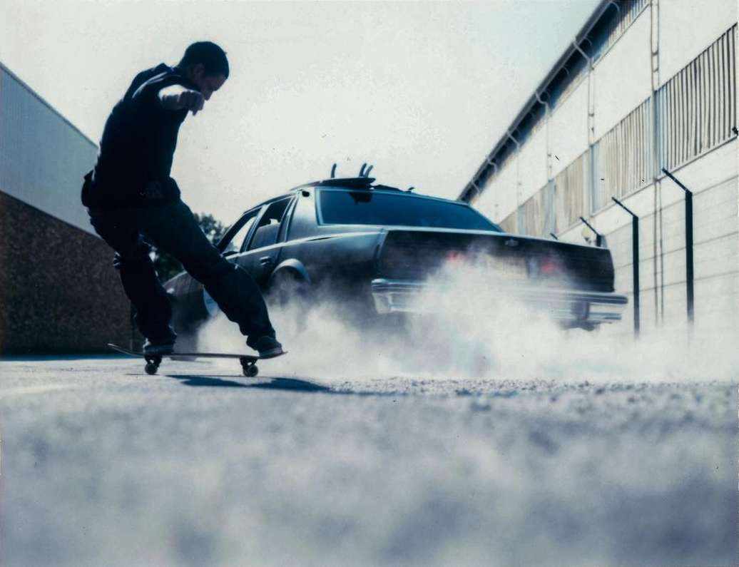 Man on a skateboard in front of a car with smoke. The photograph is taken at a low angle outside near an industrial area.