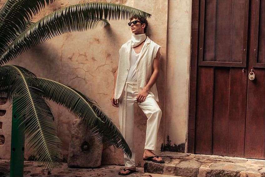 A casually dressed man leans against a weathered wall next to a dark brown door, with palm fronds framing the scene.
