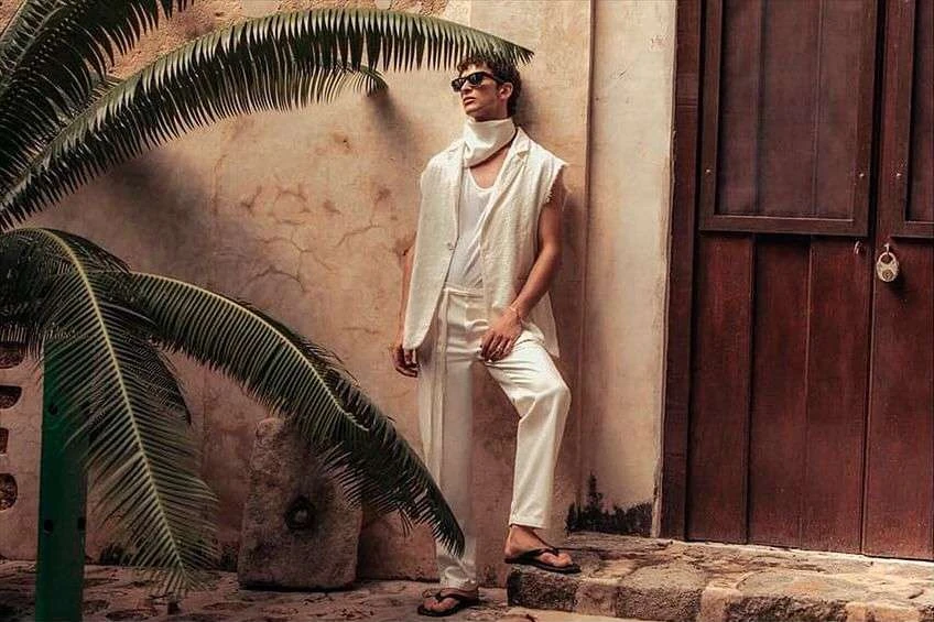 A casually dressed man leans against a weathered wall next to a dark brown door, with palm fronds framing the scene.