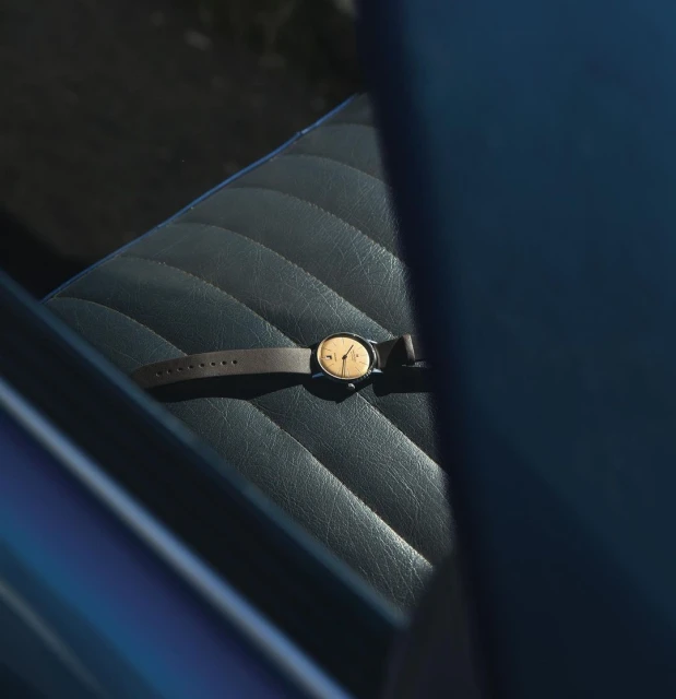 Wristwatch with a brown leather band laying on a quilted leather seat. The watch face has a golden color under direct light.
