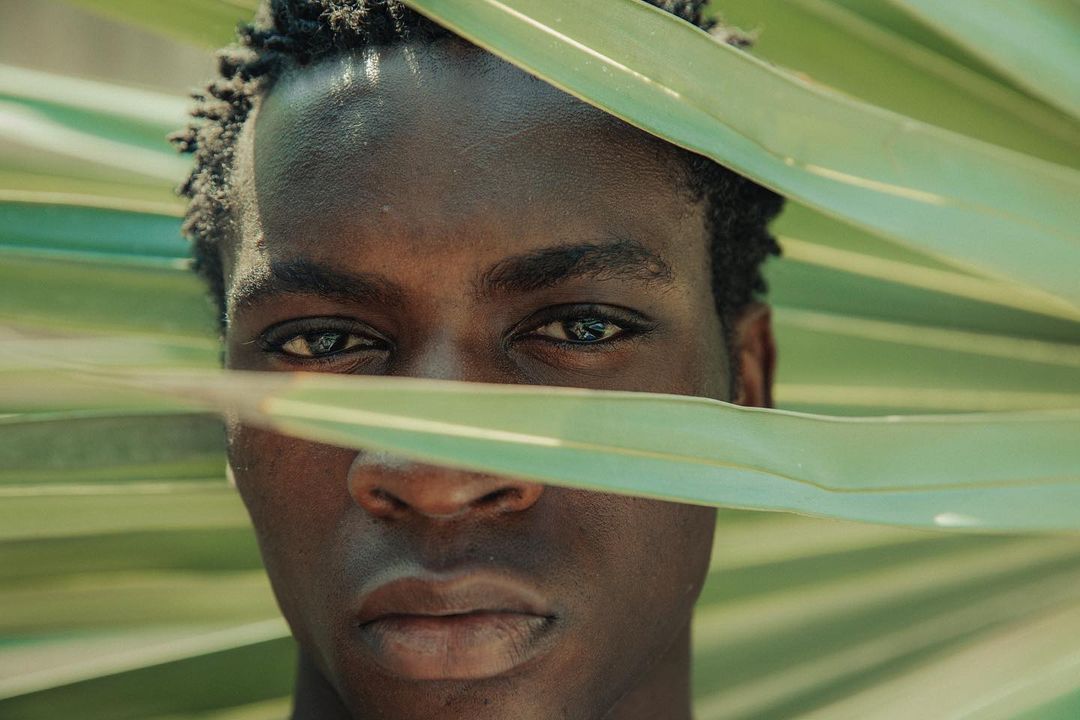 Close-up portrait of a Black man with dark hair, looking directly at the camera, partially obscured by large, light-green palm leaves.