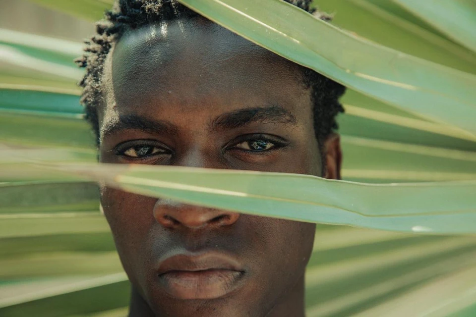 Close-up portrait of a Black man with dark hair, looking directly at the camera, partially obscured by large, light-green palm leaves.