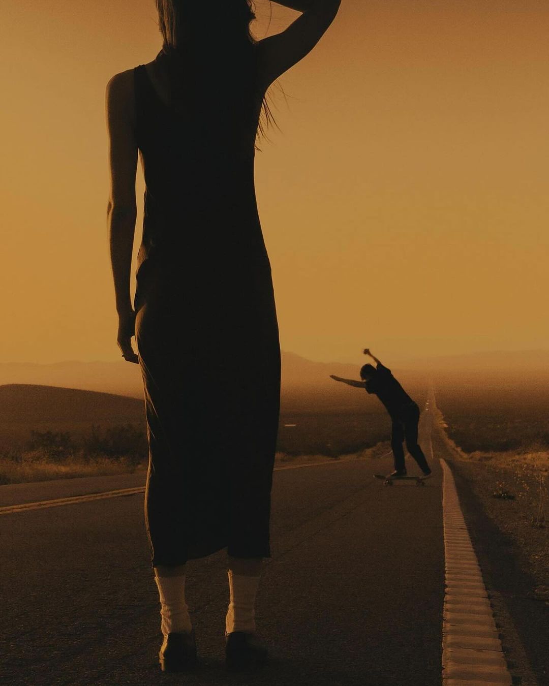 Woman in dress stands on road in hazy sunlight, while a skateboarder approaches in the distance under a clear yellow sky at golden hour.