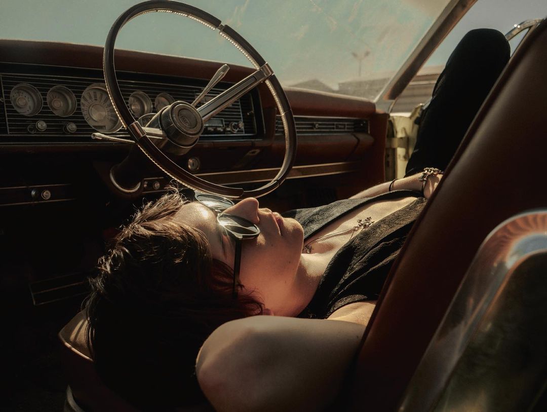 Adult laying back in the driver's seat of a classic car with a steering wheel, dashboard, and outdoor background during daylight.